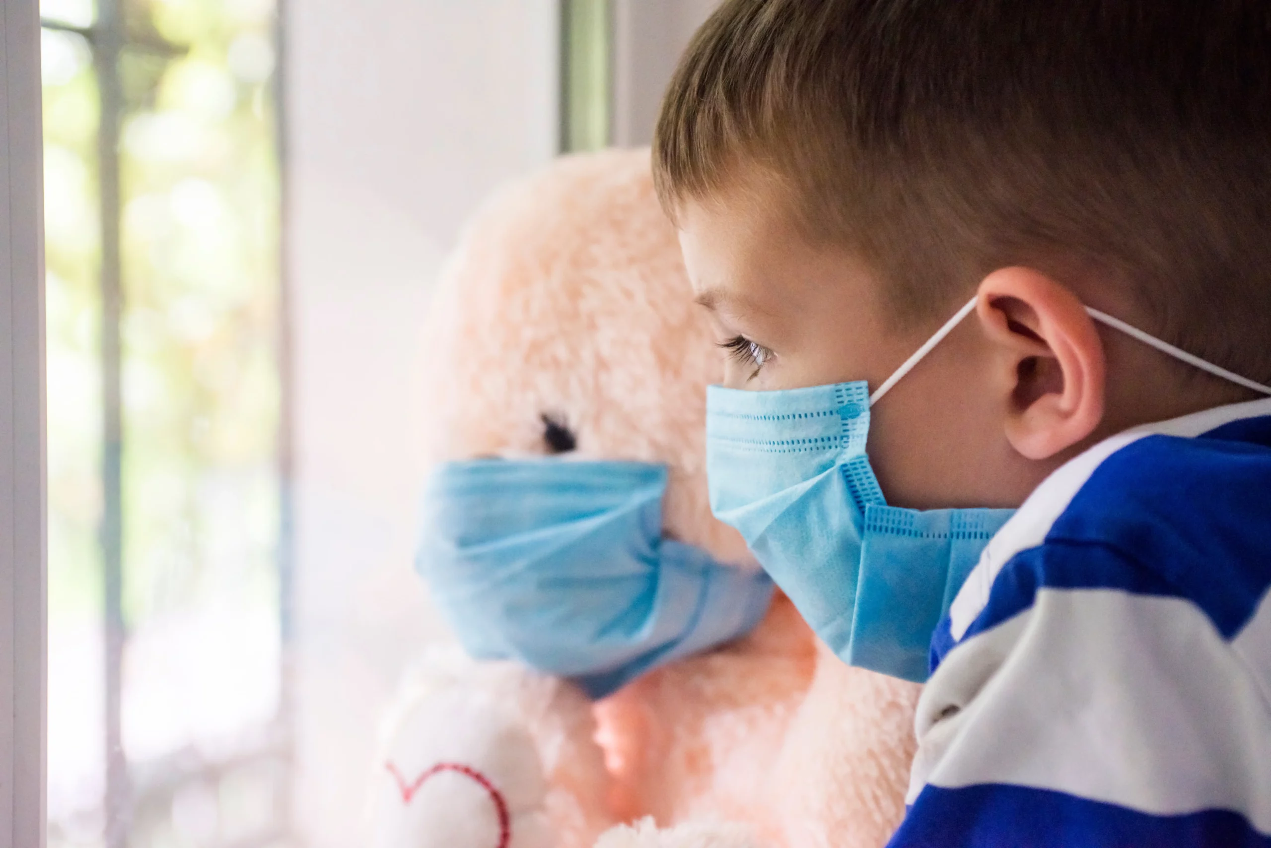 Little boy and his teddy bear holding mask and looking out of window.
